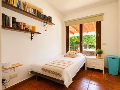 Dormitorio individual con suelos de barro artesanal, paredes blancas y acceso directo a una terraza cubierta a través de una gran puerta de cristal con marco de madera. Incluye estantes con libros.