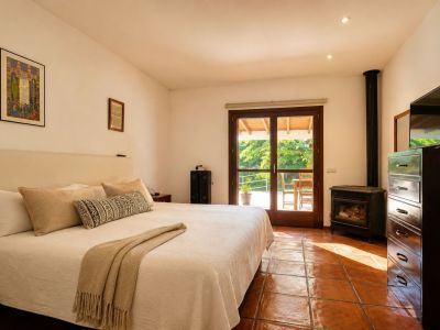 Dormitorio luminoso con cama grande, suelos de barro artesanal y una estufa de leña. La puerta de cristal doble ofrece acceso directo a una terraza exterior rodeada de vegetación.