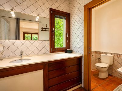 Lavabo con encimera clara y mueble de madera oscura. El gran espejo con apliques ilumina la zona, mientras una ventana de madera ofrece vistas a la vegetación exterior.