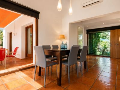 Luminoso comedor con mesa de madera oscura y seis sillas grises, sobre suelos de barro artesanal. Grandes puertas de cristal conectan con una terraza exterior bajo toldo naranja y vistas al jardín.