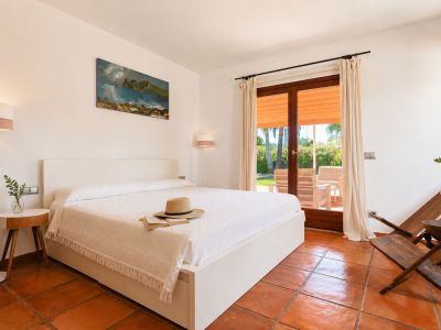 Luminoso dormitorio con cama blanca, suelos de barro artesanal y acceso directo a una terraza exterior con vistas al jardín y mobiliario.