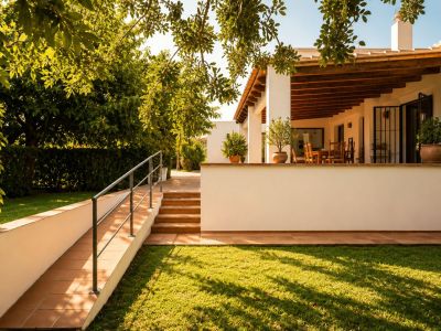 Naya cubierta con vigas de madera a la vista y comedor exterior, con vistas a un césped cuidado y jardín mediterráneo, accesible por rampa y escaleras de barro.