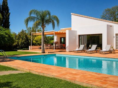 Piscina de azulejos azules rodeada de césped y baldosas de barro, con tumbonas junto a la fachada blanca de la vivienda principal y una pérgola sombreada con zona lounge.
