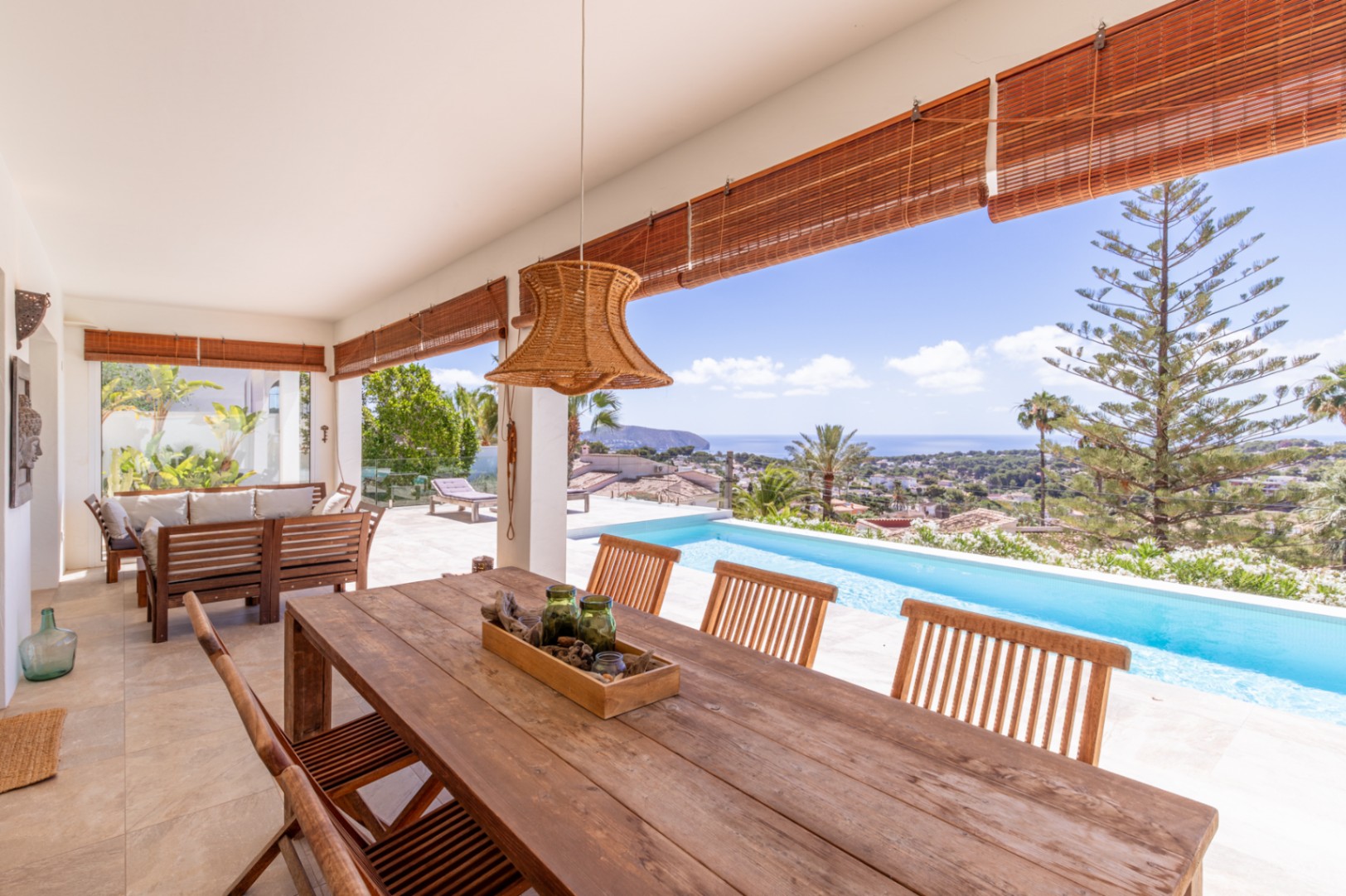 Amplia terraza cubierta con mesa de comedor de madera, lámpara de ratán y vistas panorámicas al mar y la piscina infinita. Amplia terraza cubierta con mesa de comedor de madera, lámpara de ratán y vistas panorámicas al mar y la piscina infinita.