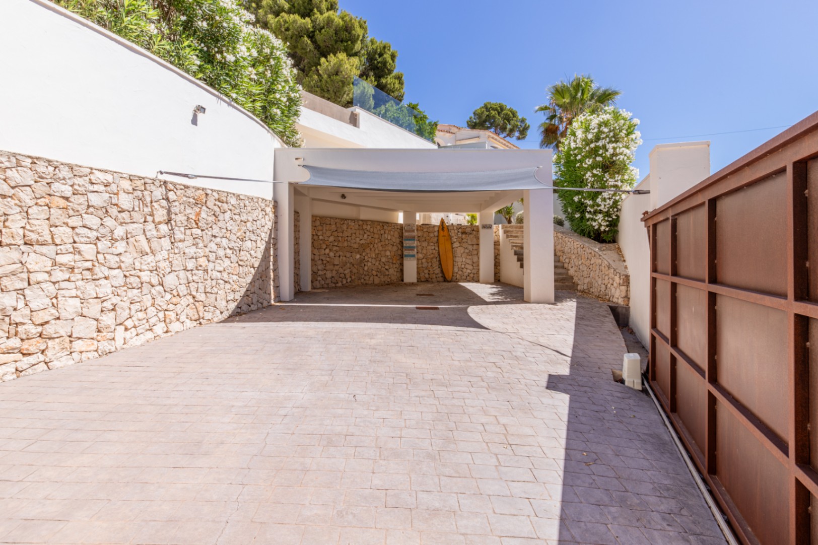 Amplio aparcamiento exterior con carport y pavimento estampado. Destaca el muro de piedra natural y la vegetación mediterránea, junto a la puerta de acceso metálica. Amplio aparcamiento exterior con carport y pavimento estampado. Destaca el muro de piedra natural y la vegetación mediterránea, junto a la puerta de acceso metálica.