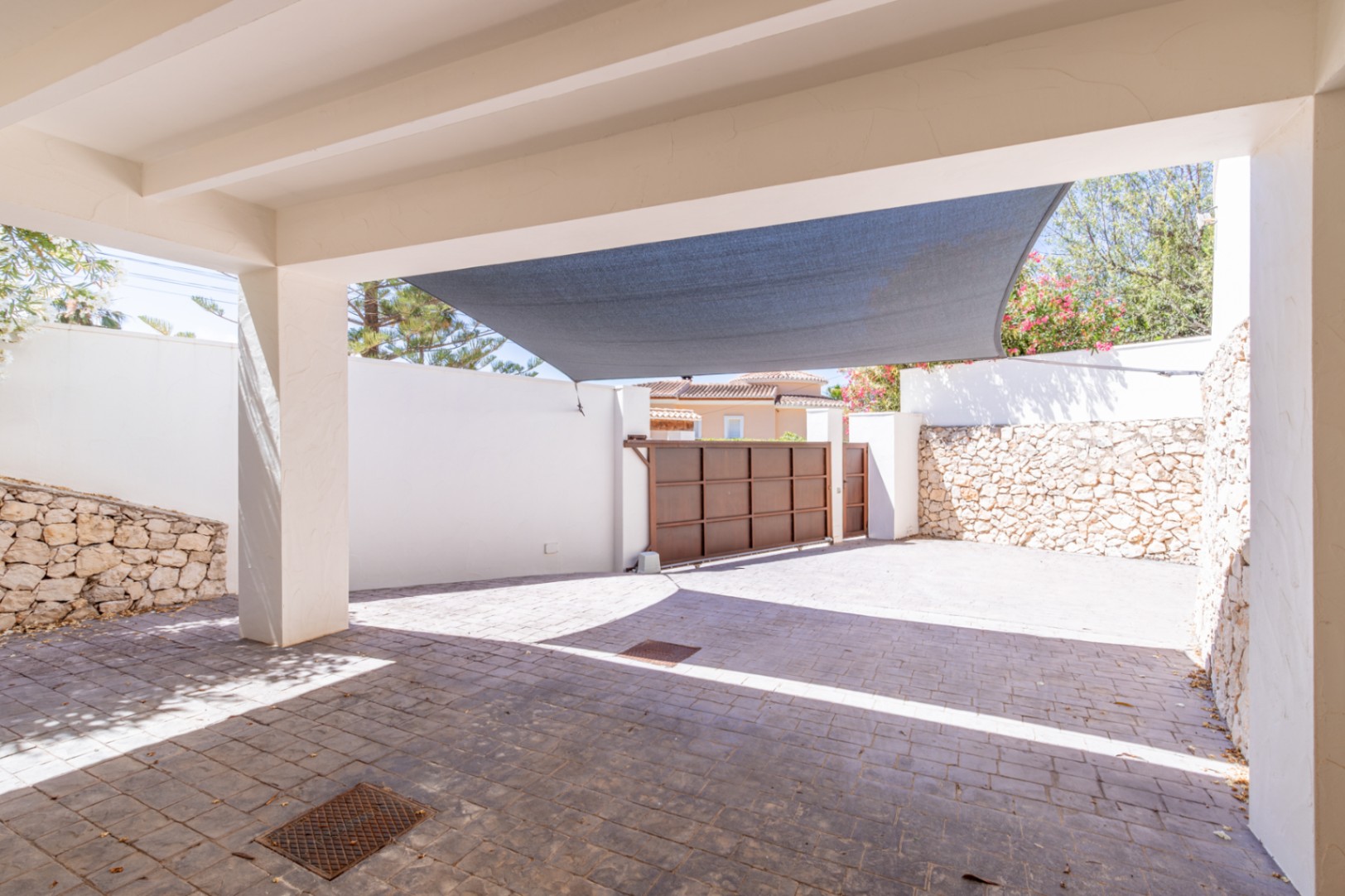 Amplio aparcamiento privado con pavimento texturizado, toldo protector y portón metálico corredero, enmarcado por muros blancos y de piedra natural. Amplio aparcamiento privado con pavimento texturizado, toldo protector y portón metálico corredero, enmarcado por muros blancos y de piedra natural.