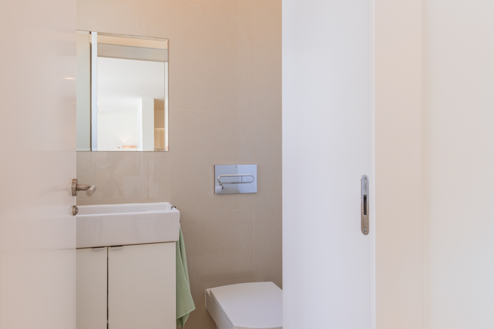 Aseo de cortesía moderno con lavabo blanco sobre mueble, espejo, inodoro suspendido y paredes de azulejos beige. Diseño funcional y limpio. Aseo de cortesía moderno con lavabo blanco sobre mueble, espejo, inodoro suspendido y paredes de azulejos beige. Diseño funcional y limpio.