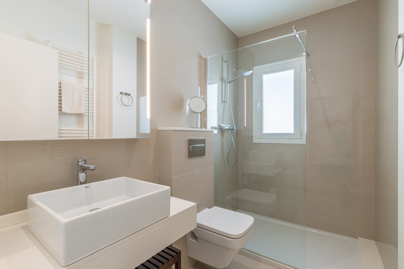 Baño moderno con lavabo rectangular sobre encimera, inodoro suspendido y ducha con mampara de cristal. Destacan los azulejos neutros y el espejo con iluminación LED integrada. Baño moderno con lavabo rectangular sobre encimera, inodoro suspendido y ducha con mampara de cristal. Destacan los azulejos neutros y el espejo con iluminación LED integrada.