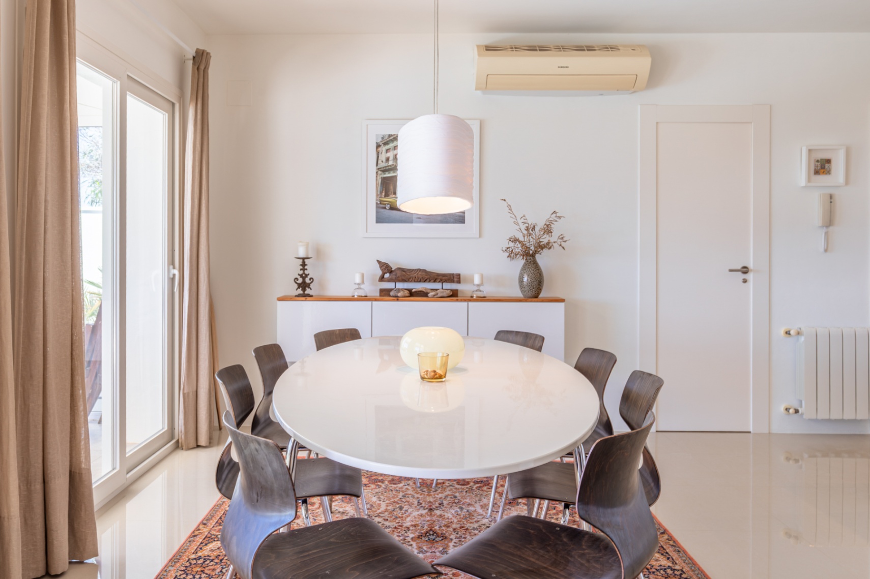 Comedor luminoso con mesa ovalada blanca y sillas de madera oscura sobre alfombra. Puerta corredera acristalada con cortinas y aparador decorado. Comedor luminoso con mesa ovalada blanca y sillas de madera oscura sobre alfombra. Puerta corredera acristalada con cortinas y aparador decorado.