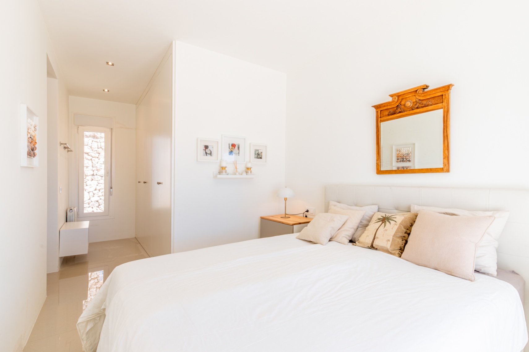 Dormitorio luminoso con cama blanca, cabecero acolchado y mesita de noche de madera. Un espejo ornamental y un pasillo con ventana estrecha hacia una pared de piedra completan la estancia. Dormitorio luminoso con cama blanca, cabecero acolchado y mesita de noche de madera. Un espejo ornamental y un pasillo con ventana estrecha hacia una pared de piedra completan la estancia.
