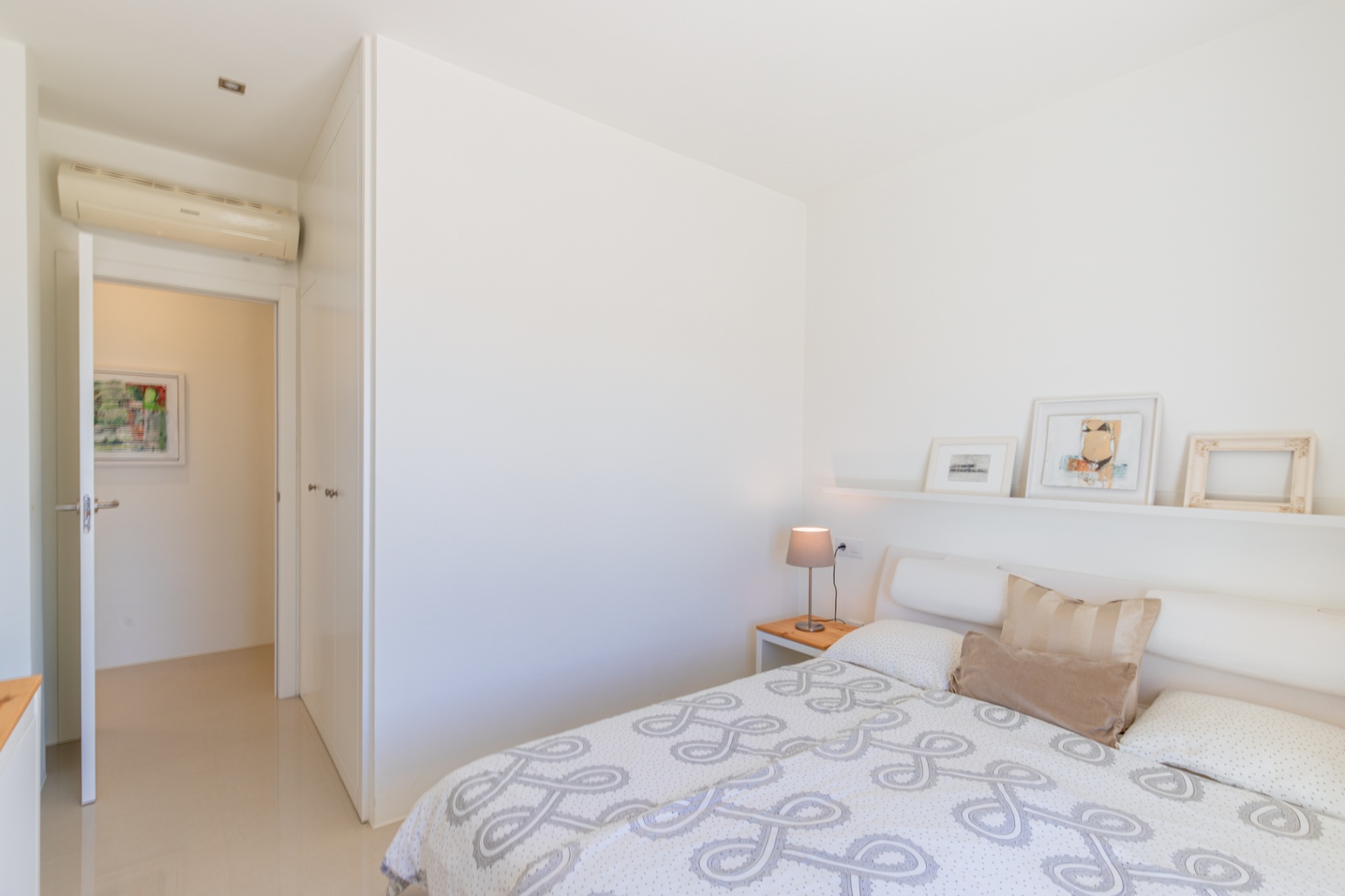 Dormitorio moderno con paredes blancas, suelo de azulejos claros y cama con edredón estampado. Dispone de mesita de noche de madera, estante decorativo, armarios empotrados y aire acondicionado. Dormitorio moderno con paredes blancas, suelo de azulejos claros y cama con edredón estampado. Dispone de mesita de noche de madera, estante decorativo, armarios empotrados y aire acondicionado.