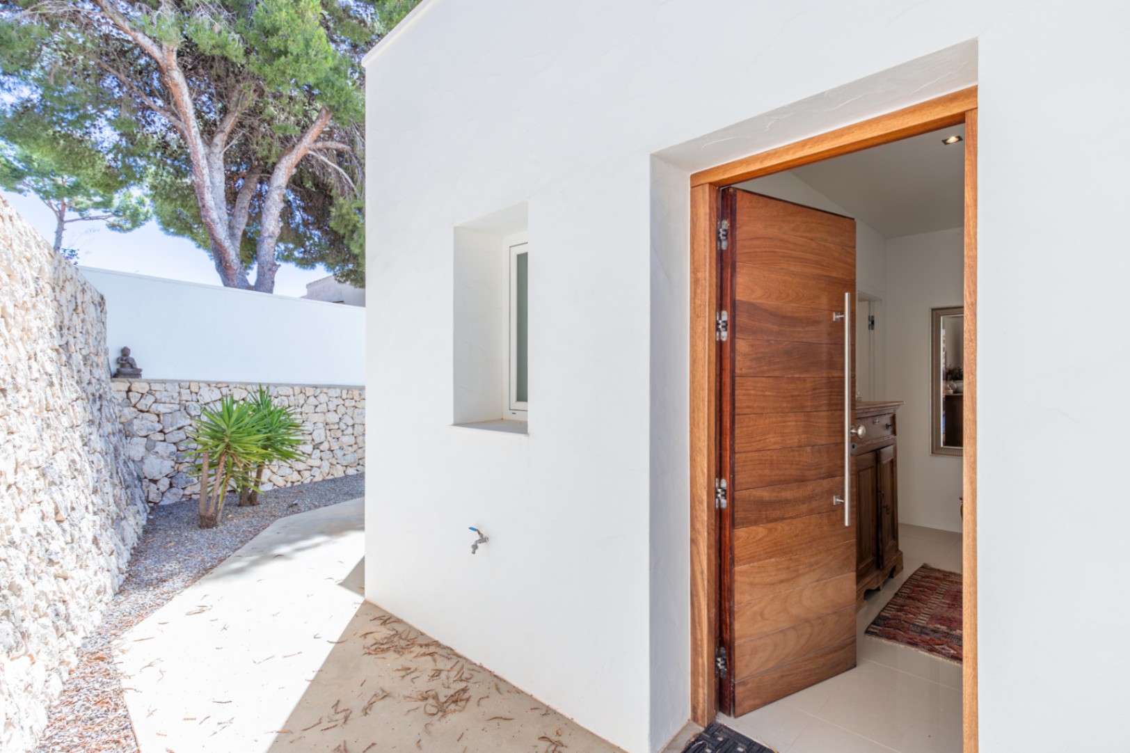Entrada principal con puerta de madera maciza y diseño moderno, enmarcada por paredes blancas y un muro de piedra seca. Un camino de hormigón conduce al interior, revelando un recibidor luminoso. Entrada principal con puerta de madera maciza y diseño moderno, enmarcada por paredes blancas y un muro de piedra seca. Un camino de hormigón conduce al interior, revelando un recibidor luminoso.