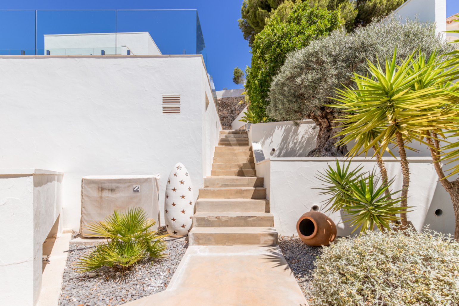 Escalera exterior con paredes blancas de estuco, plantas mediterráneas y paisajismo de grava, que conduce a un nivel superior bajo un cielo azul claro. Escalera exterior con paredes blancas de estuco, plantas mediterráneas y paisajismo de grava, que conduce a un nivel superior bajo un cielo azul claro.