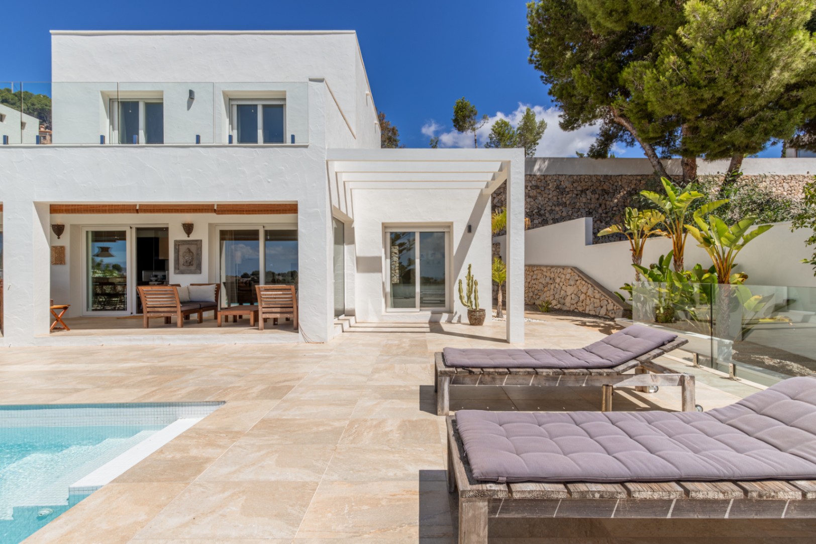 Exterior de villa blanca moderna con piscina de azulejos claros, terraza soleada con tumbonas de madera y un área de estar cubierta con muebles de madera, enmarcada por un jardín escalonado con muros de piedra. Exterior de villa blanca moderna con piscina de azulejos claros, terraza soleada con tumbonas de madera y un área de estar cubierta con muebles de madera, enmarcada por un jardín escalonado con muros de piedra.