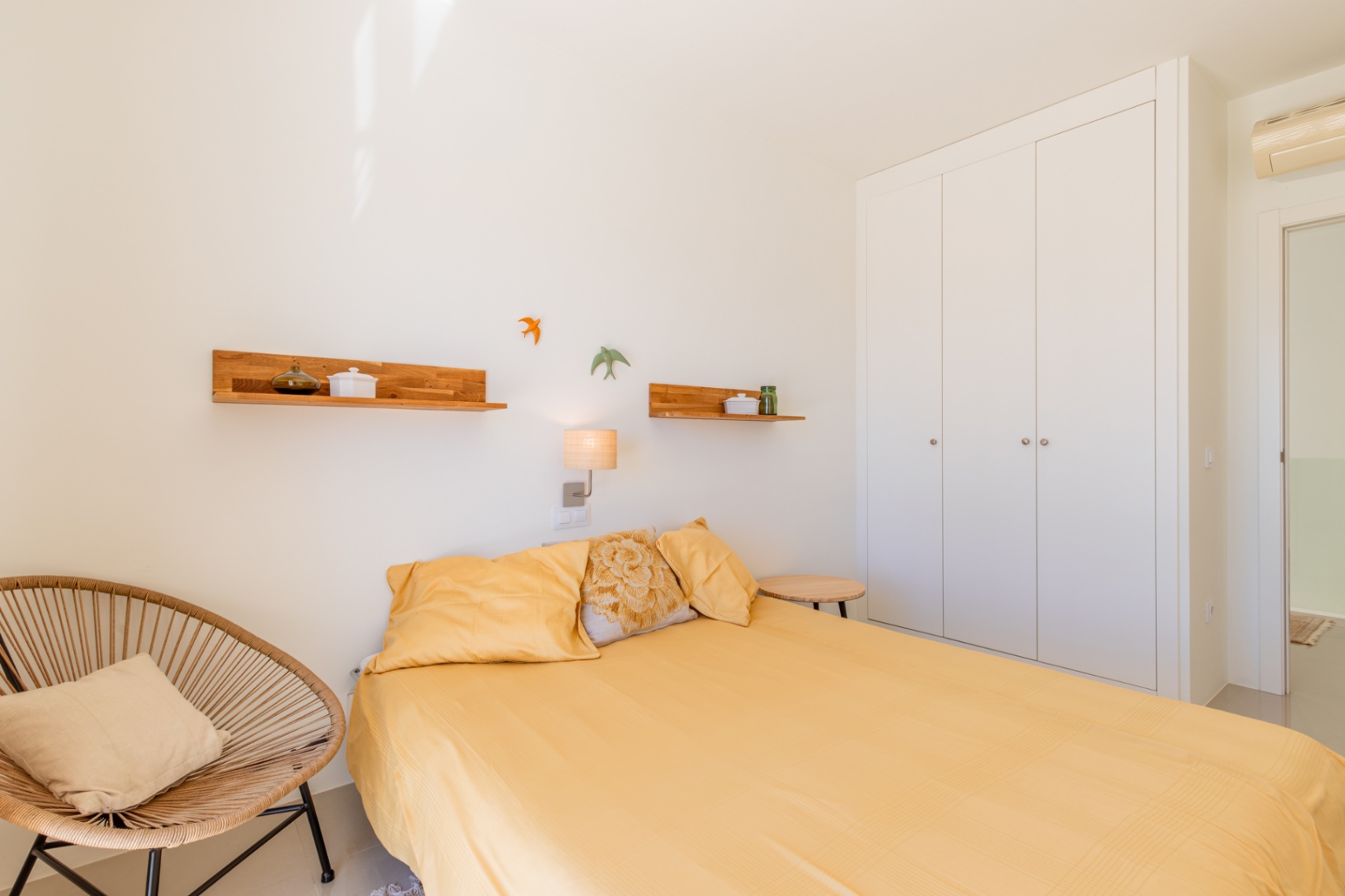 Luminoso dormitorio con cama de sábanas amarillas, armario empotrado blanco y un sillón de mimbre. Estantes de madera y una lámpara de pared complementan el espacio. Luminoso dormitorio con cama de sábanas amarillas, armario empotrado blanco y un sillón de mimbre. Estantes de madera y una lámpara de pared complementan el espacio.