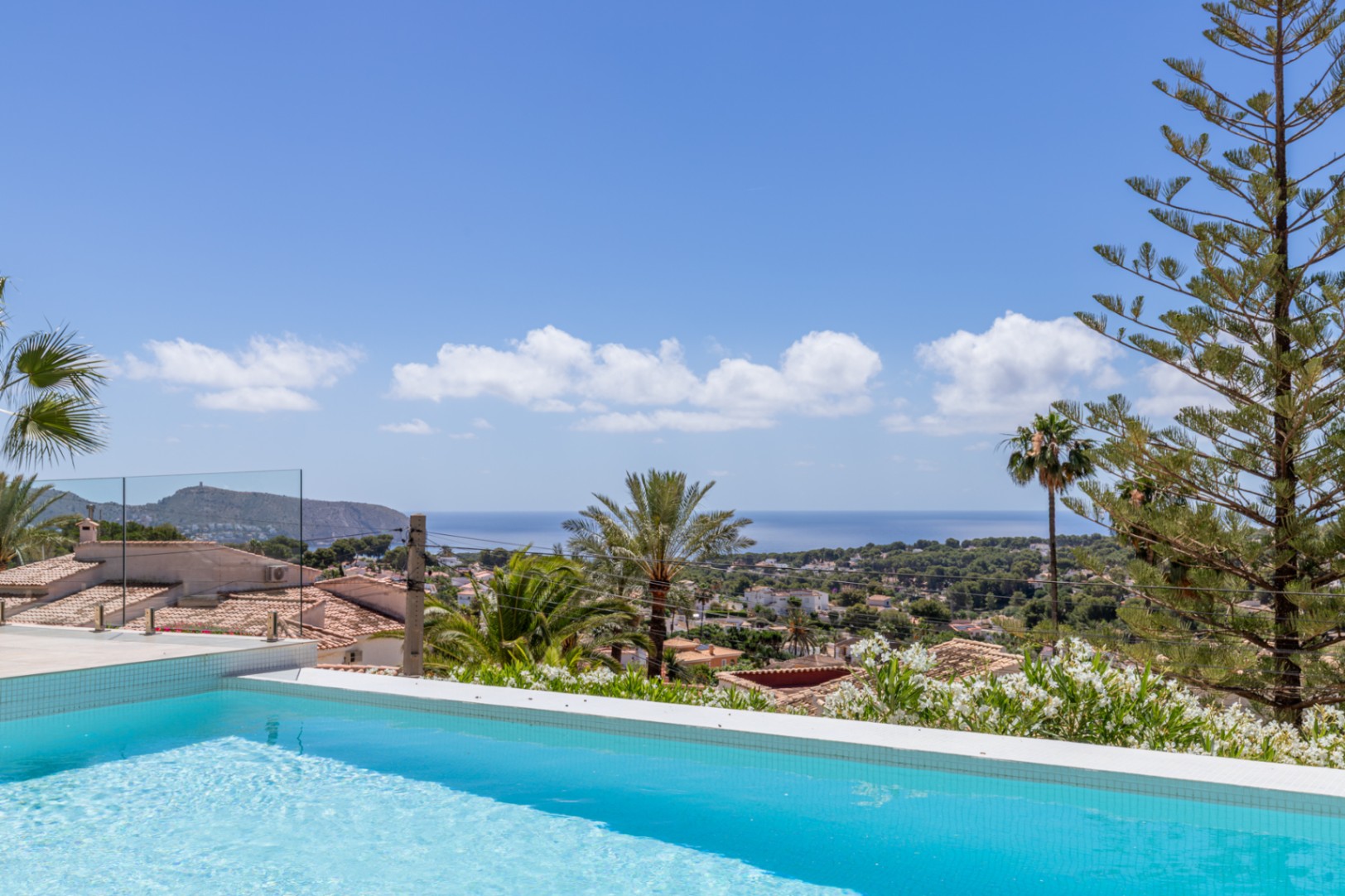 Piscina de azulejos azules con vistas panorámicas al Mediterráneo, tejados tradicionales y vegetación exuberante bajo un cielo despejado. Barandilla de cristal. Piscina de azulejos azules con vistas panorámicas al Mediterráneo, tejados tradicionales y vegetación exuberante bajo un cielo despejado. Barandilla de cristal.