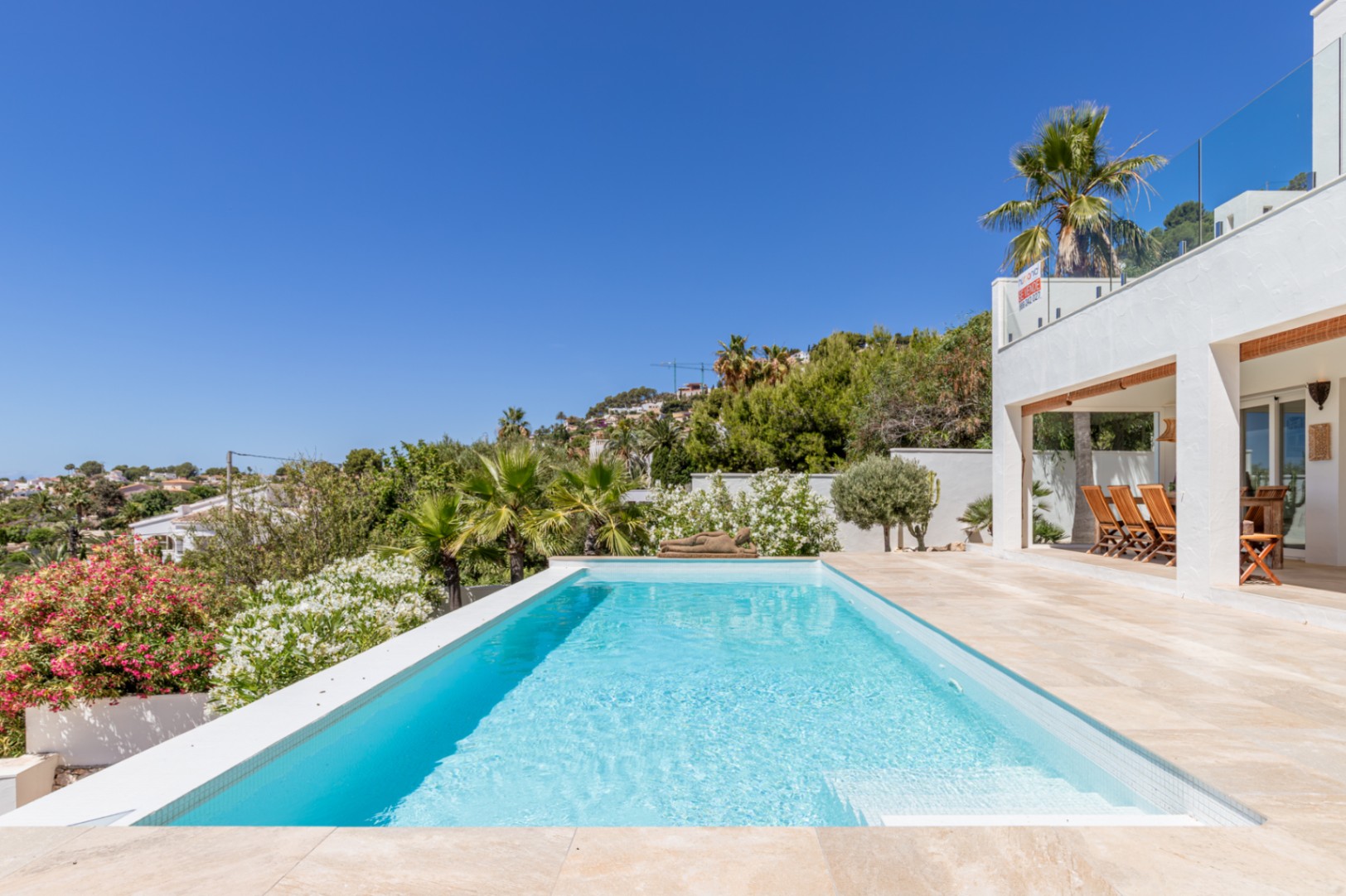 Piscina rectangular de agua azul claro con borde infinito, rodeada de una terraza de gres y exuberante vegetación mediterránea. Se aprecia una zona de comedor exterior cubierta y la fachada blanca de la villa. Piscina rectangular de agua azul claro con borde infinito, rodeada de una terraza de gres y exuberante vegetación mediterránea. Se aprecia una zona de comedor exterior cubierta y la fachada blanca de la villa.