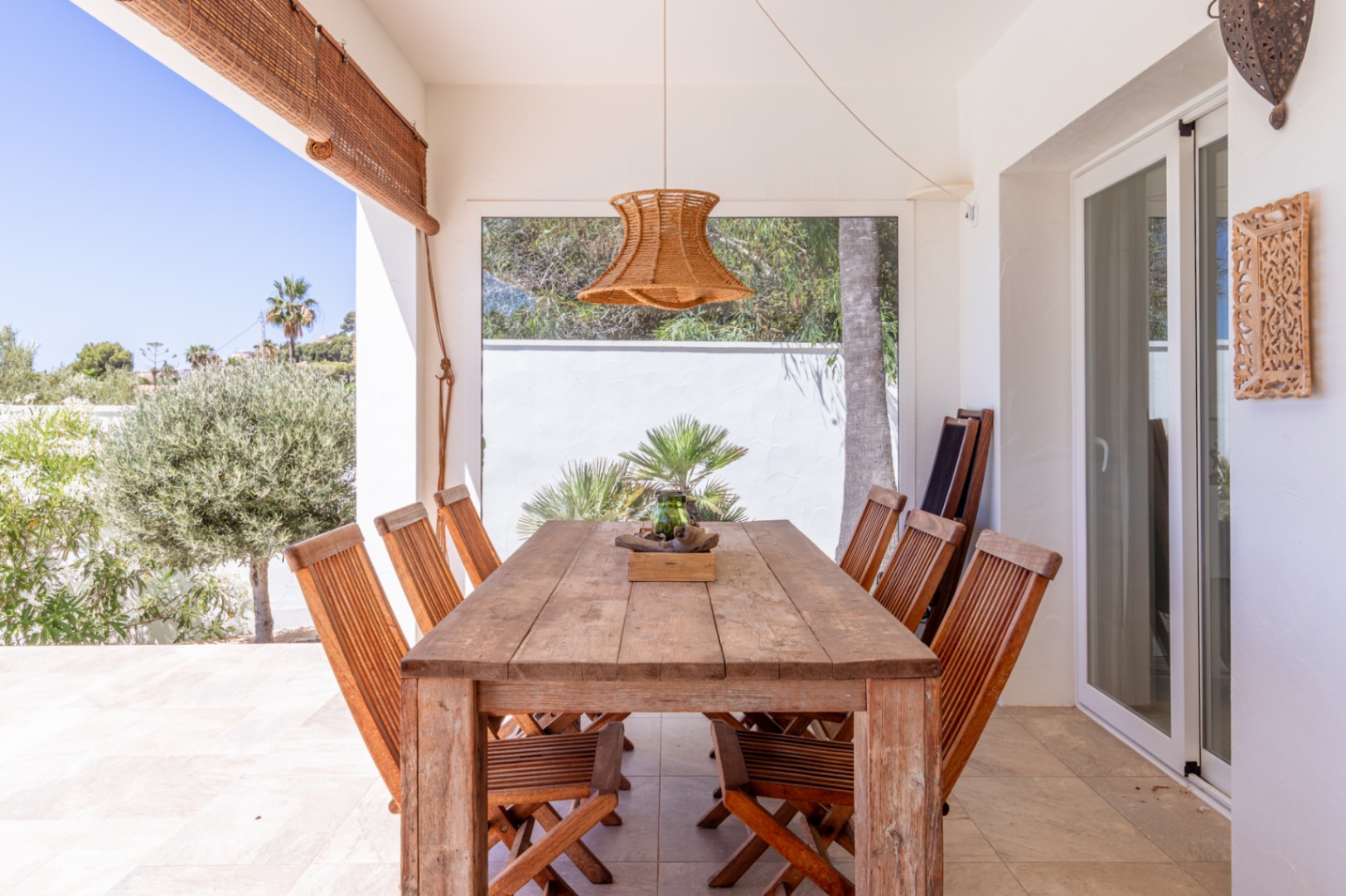Terraza cubierta con mesa y sillas de madera rústica, bajo una lámpara colgante de fibra natural. Vistas directas a la exuberante vegetación mediterránea. Terraza cubierta con mesa y sillas de madera rústica, bajo una lámpara colgante de fibra natural. Vistas directas a la exuberante vegetación mediterránea.