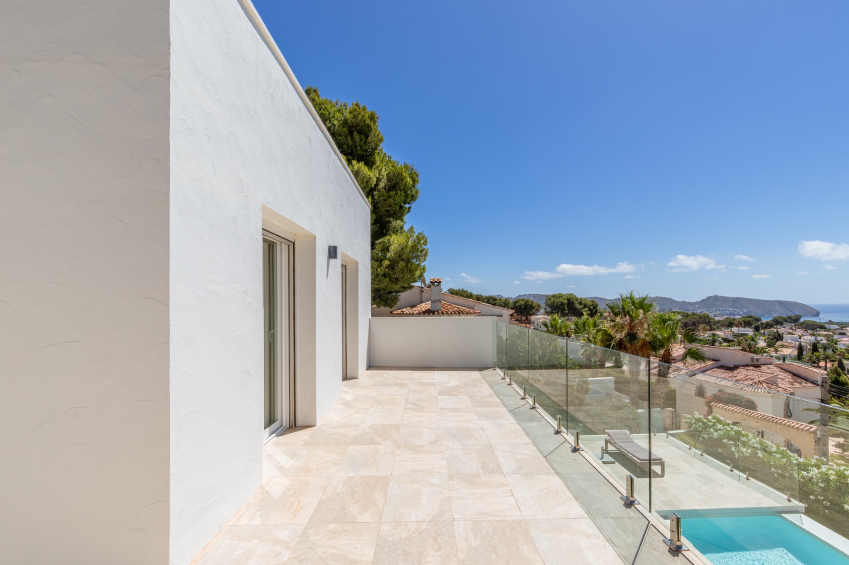 Terraza superior con suelo de baldosas claras y barandilla de cristal, ofreciendo vistas panorámicas al Mediterráneo y la costa. Se aprecian puertas acristaladas y parte de una piscina inferior. Terraza superior con suelo de baldosas claras y barandilla de cristal, ofreciendo vistas panorámicas al Mediterráneo y la costa. Se aprecian puertas acristaladas y parte de una piscina inferior.