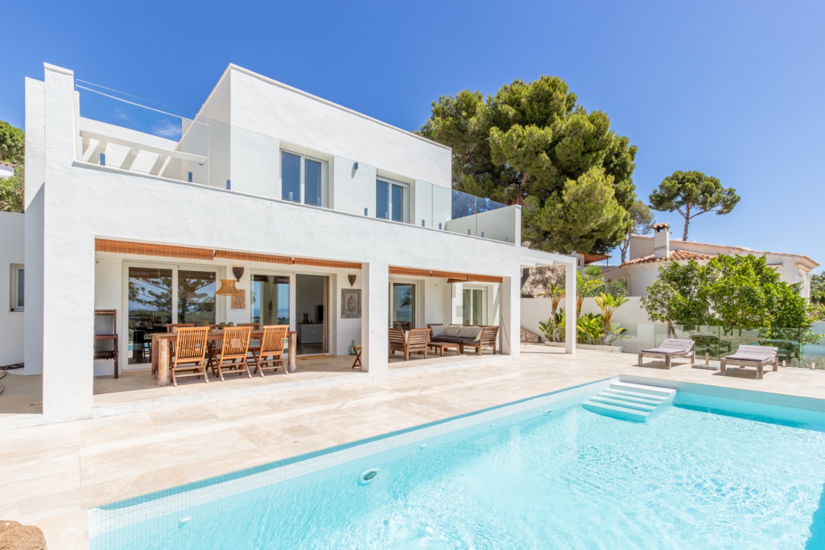 Villa moderna con piscina de azulejos claros y amplias terrazas. Cuenta con comedor exterior cubierto, zona lounge y barandillas de cristal en la planta superior. Villa moderna con piscina de azulejos claros y amplias terrazas. Cuenta con comedor exterior cubierto, zona lounge y barandillas de cristal en la planta superior.