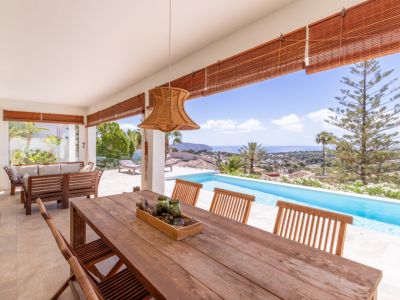 Amplia terraza cubierta con mesa de comedor de madera, lámpara de ratán y vistas panorámicas al mar y la piscina infinita.