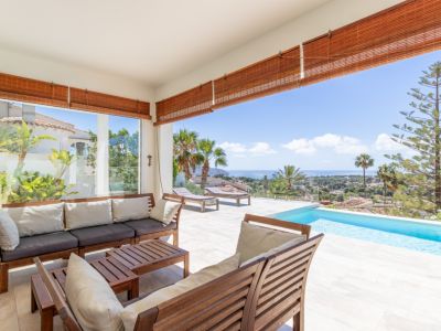 Amplia terraza cubierta con mobiliario de madera, suelo de baldosas claras y piscina infinita, ofreciendo vistas panorámicas al mar Mediterráneo.