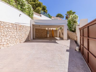 Amplio aparcamiento exterior con carport y pavimento estampado. Destaca el muro de piedra natural y la vegetación mediterránea, junto a la puerta de acceso metálica.