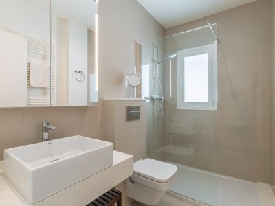 Baño moderno con lavabo rectangular sobre encimera, inodoro suspendido y ducha con mampara de cristal. Destacan los azulejos neutros y el espejo con iluminación LED integrada.