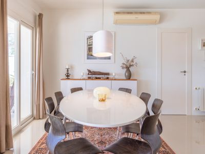 Comedor luminoso con mesa ovalada blanca y sillas de madera oscura sobre alfombra. Puerta corredera acristalada con cortinas y aparador decorado.