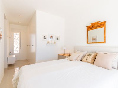 Dormitorio luminoso con cama blanca, cabecero acolchado y mesita de noche de madera. Un espejo ornamental y un pasillo con ventana estrecha hacia una pared de piedra completan la estancia.