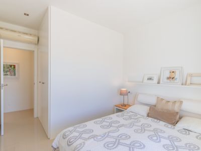 Dormitorio moderno con paredes blancas, suelo de azulejos claros y cama con edredón estampado. Dispone de mesita de noche de madera, estante decorativo, armarios empotrados y aire acondicionado.