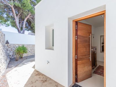 Entrada principal con puerta de madera maciza y diseño moderno, enmarcada por paredes blancas y un muro de piedra seca. Un camino de hormigón conduce al interior, revelando un recibidor luminoso.