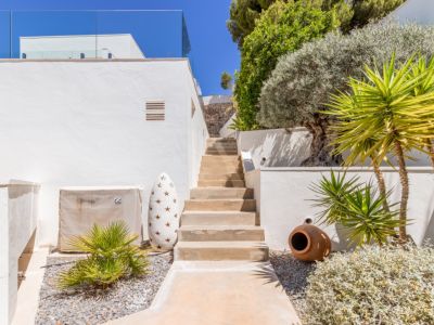Escalera exterior con paredes blancas de estuco, plantas mediterráneas y paisajismo de grava, que conduce a un nivel superior bajo un cielo azul claro.