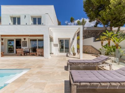 Exterior de villa blanca moderna con piscina de azulejos claros, terraza soleada con tumbonas de madera y un área de estar cubierta con muebles de madera, enmarcada por un jardín escalonado con muros de piedra.