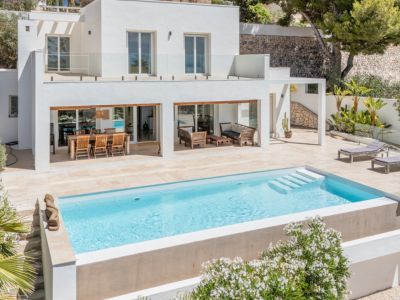 La villa moderna exhibe una piscina rectangular con escalones y una amplia terraza de piedra clara. Incluye zona de comedor exterior y salón, con balcón superior de barandilla de cristal.