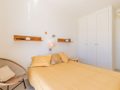 Luminoso dormitorio con cama de sábanas amarillas, armario empotrado blanco y un sillón de mimbre. Estantes de madera y una lámpara de pared complementan el espacio.