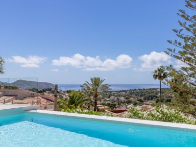Piscina de azulejos azules con vistas panorámicas al Mediterráneo, tejados tradicionales y vegetación exuberante bajo un cielo despejado. Barandilla de cristal.