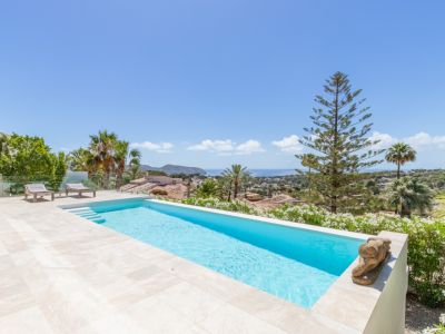 Piscina infinita con baldosas claras y terraza de gres, ofreciendo vistas panorámicas ininterrumpidas al Mediterráneo. Dos tumbonas y exuberante vegetación costera completan el exterior.