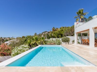 Piscina rectangular de agua azul claro con borde infinito, rodeada de una terraza de gres y exuberante vegetación mediterránea. Se aprecia una zona de comedor exterior cubierta y la fachada blanca de la villa.
