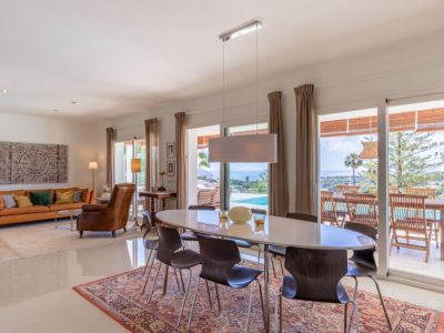 Salón-comedor de concepto abierto con mesa ovalada, sofá naranja y vistas panorámicas al Mediterráneo desde sus amplios ventanales.