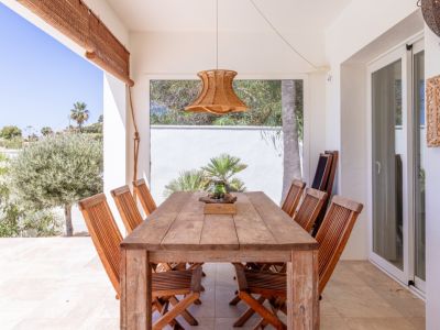 Terraza cubierta con mesa y sillas de madera rústica, bajo una lámpara colgante de fibra natural. Vistas directas a la exuberante vegetación mediterránea.