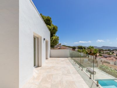 Terraza superior con suelo de baldosas claras y barandilla de cristal, ofreciendo vistas panorámicas al Mediterráneo y la costa. Se aprecian puertas acristaladas y parte de una piscina inferior.