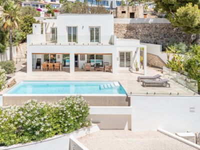 Villa moderna de dos plantas con piscina rectangular y amplia terraza solada. Destacan la zona de comedor exterior cubierta y la barandilla de cristal, integrando el diseño con el entorno mediterráneo.