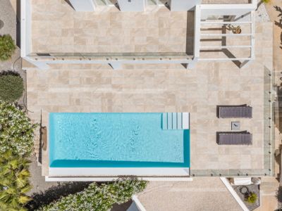 Vista aérea de la piscina rectangular de agua azul, rodeada por una amplia terraza de baldosas claras, con tumbonas y vegetación mediterránea.