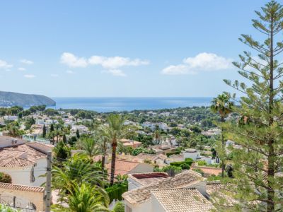 Vista elevada de la costa mediterránea, mostrando tejados de teja, exuberante vegetación y un promontorio rocoso que se adentra en el mar azul bajo un cielo despejado.