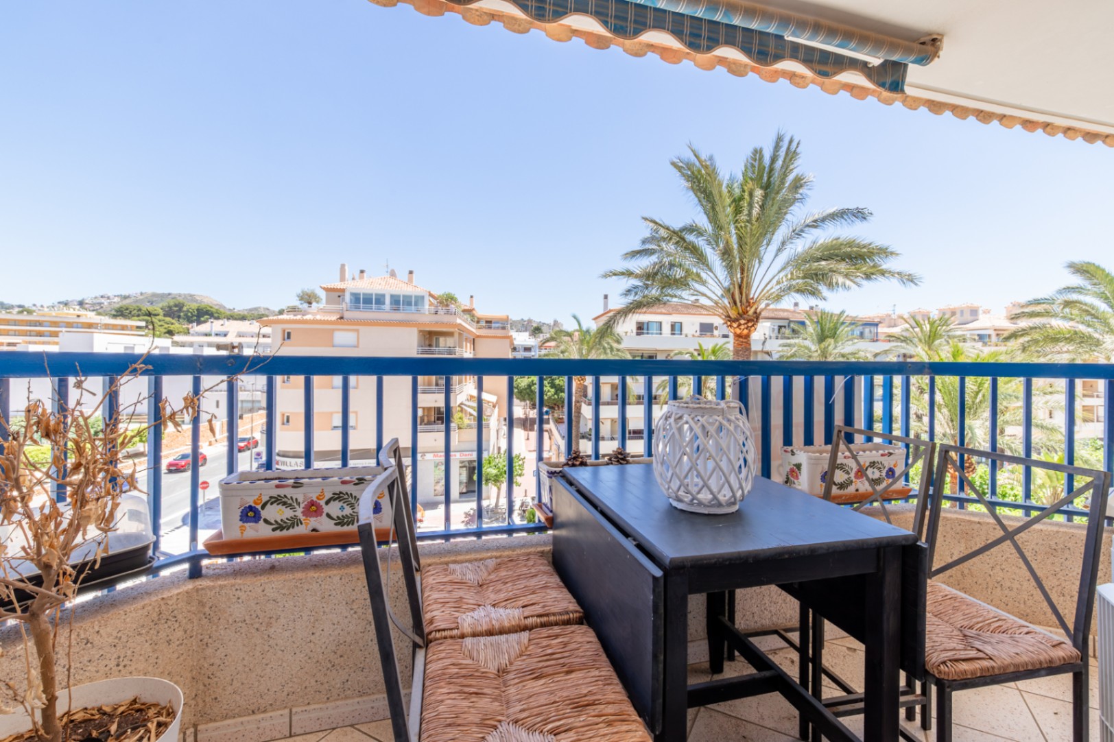 Balcón exterior con mesa plegable y sillas de mimbre, barandilla azul y vistas a la calle, edificios y palmeras bajo un toldo. Balcón exterior con mesa plegable y sillas de mimbre, barandilla azul y vistas a la calle, edificios y palmeras bajo un toldo.