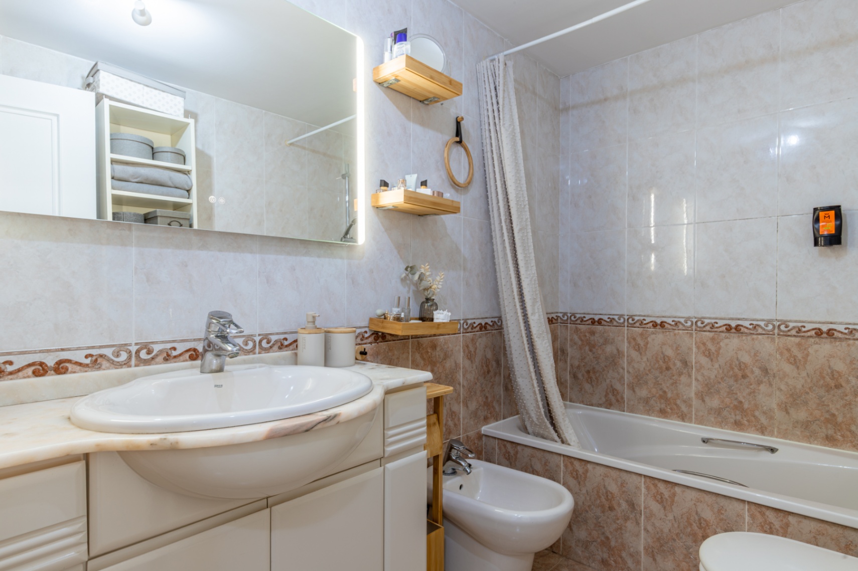 Baño completo con azulejos claros y cenefa decorativa, lavabo integrado en encimera de mármol, espejo retroiluminado, bañera con cortina y bidé. Baño completo con azulejos claros y cenefa decorativa, lavabo integrado en encimera de mármol, espejo retroiluminado, bañera con cortina y bidé.