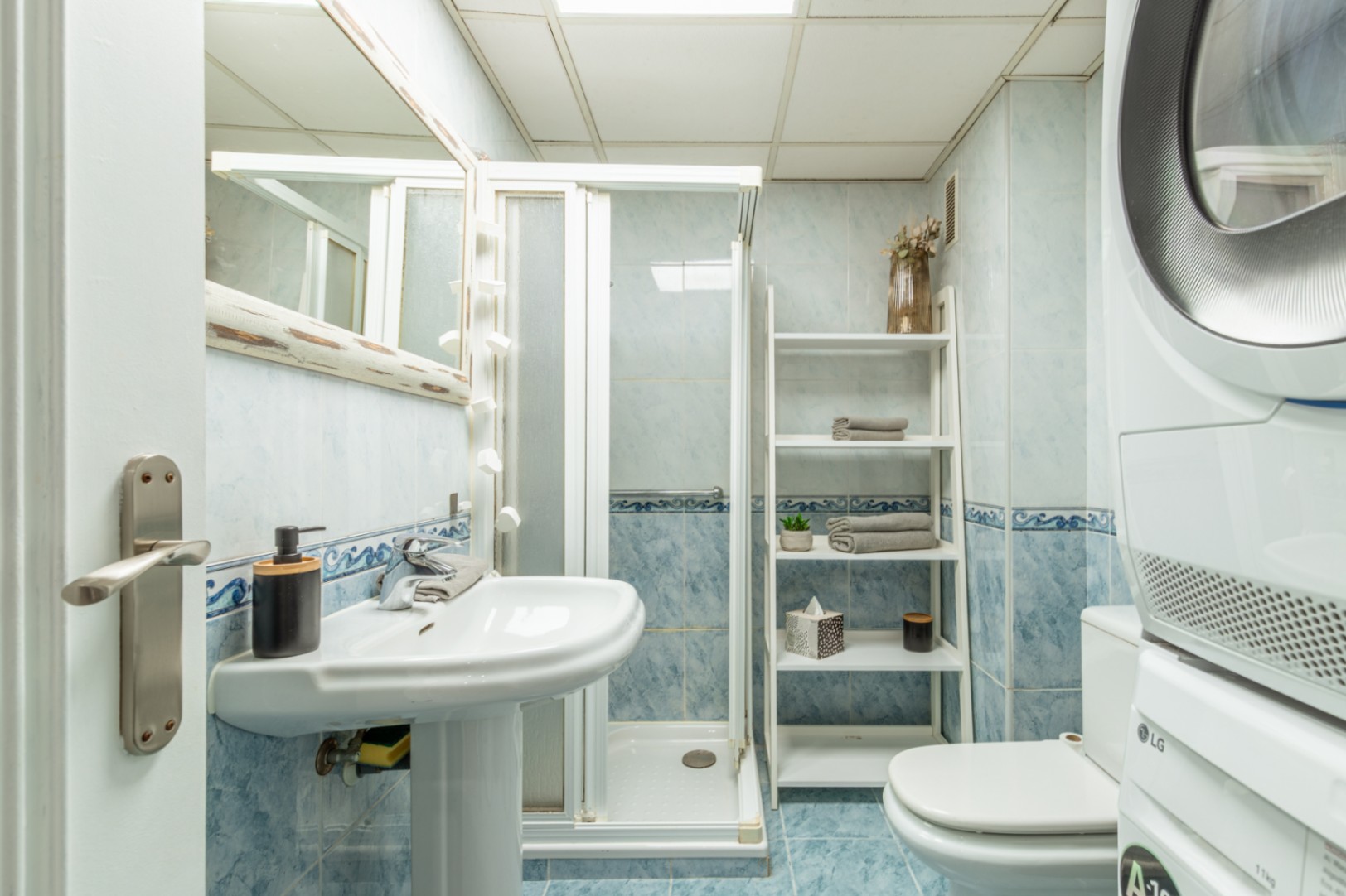 Baño funcional con azulejos azules, lavabo de pedestal, cabina de ducha y lavadora/secadora LG apiladas. Estantería tipo escalera para almacenamiento. Baño funcional con azulejos azules, lavabo de pedestal, cabina de ducha y lavadora/secadora LG apiladas. Estantería tipo escalera para almacenamiento.