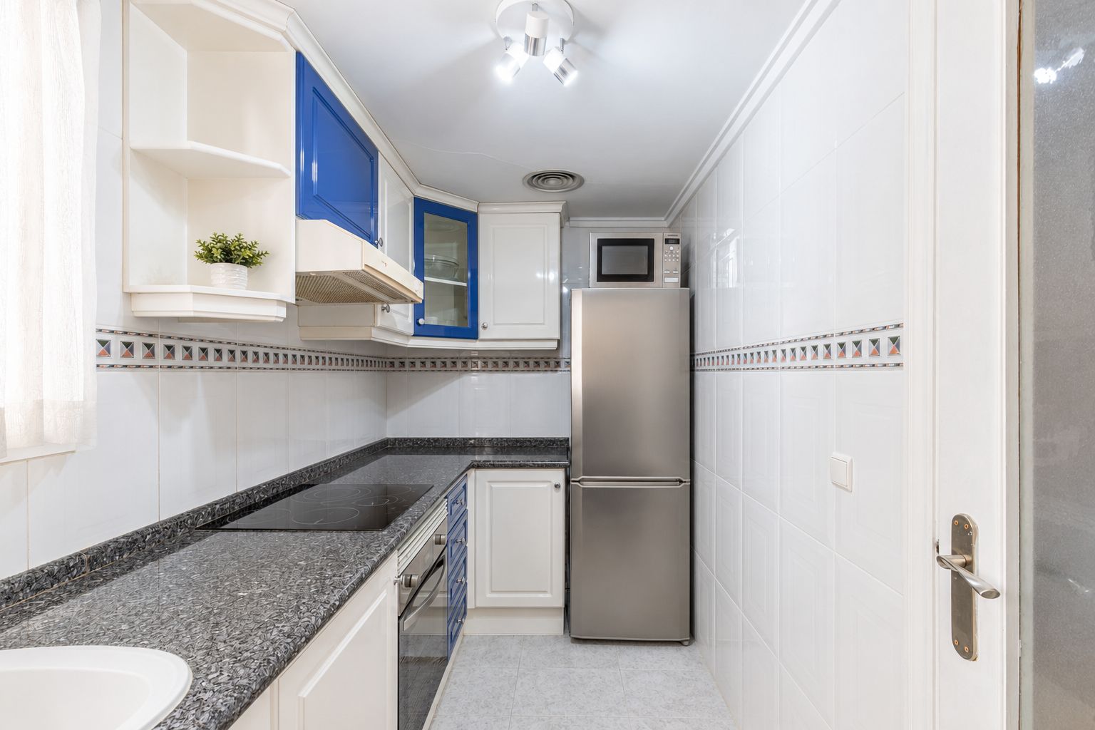 Cocina funcional con armarios blancos y azules, encimeras de granito oscuro, placa de inducción integrada y electrodomésticos de acero inoxidable. Iluminación natural junto a la ventana. Cocina funcional con armarios blancos y azules, encimeras de granito oscuro, placa de inducción integrada y electrodomésticos de acero inoxidable. Iluminación natural junto a la ventana.