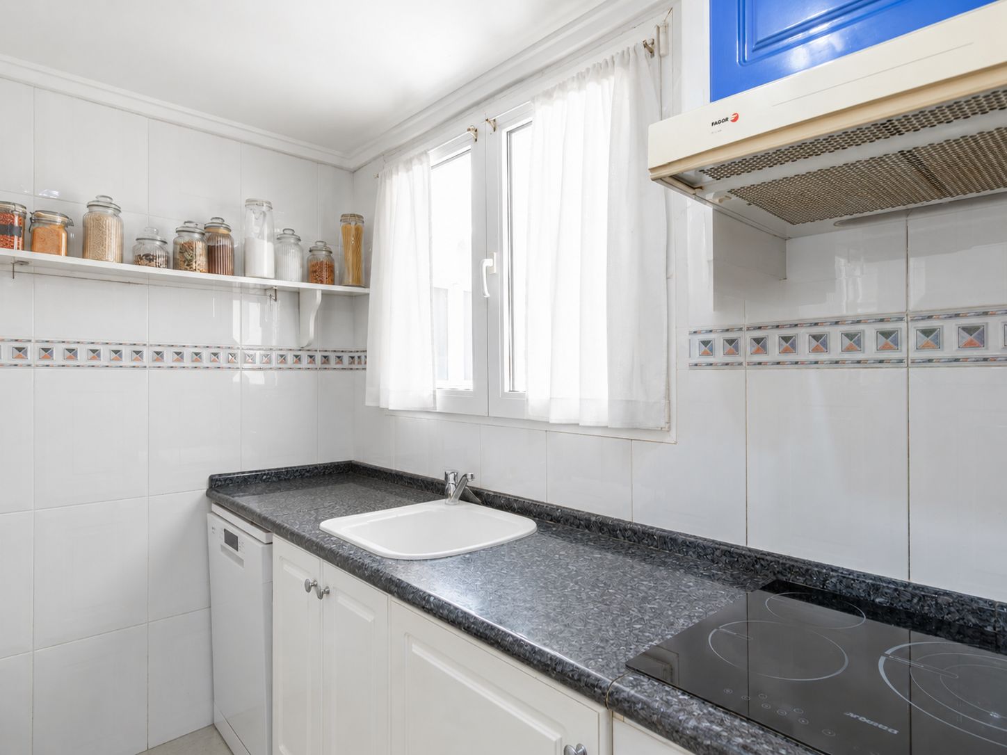 Cocina independiente con encimera de granito oscuro, fregadero blanco y placa de inducción. Paredes alicatadas en blanco con cenefa decorativa y ventana con luz natural. Cocina independiente con encimera de granito oscuro, fregadero blanco y placa de inducción. Paredes alicatadas en blanco con cenefa decorativa y ventana con luz natural.
